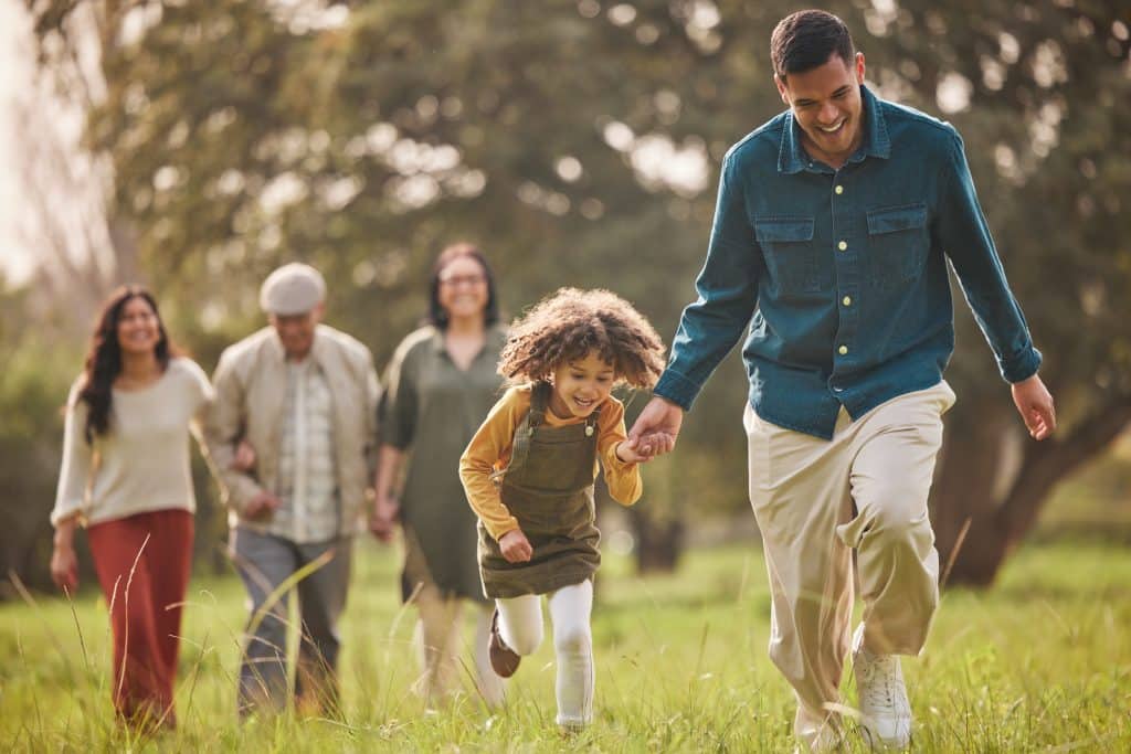 A smiling family of five walking through a grassy field, with a man and a young girl holding hands in the foreground in bozeman mt