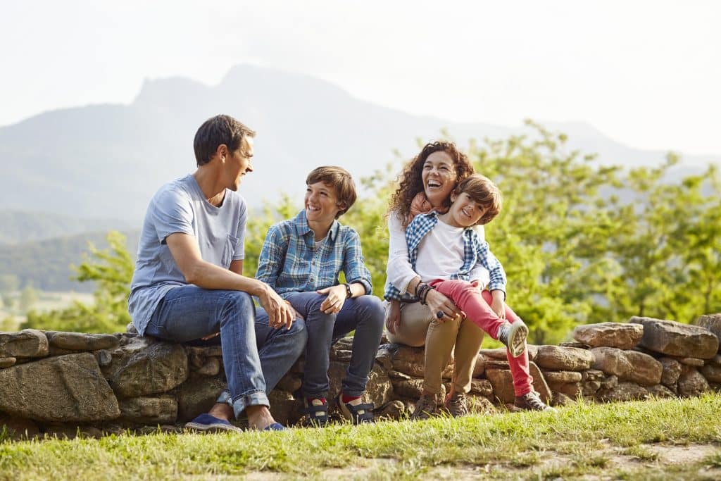 A family of four sitting on a stone wall in a grassy area, smiling and talking, with a mountainous landscape in the background in bozeman mt